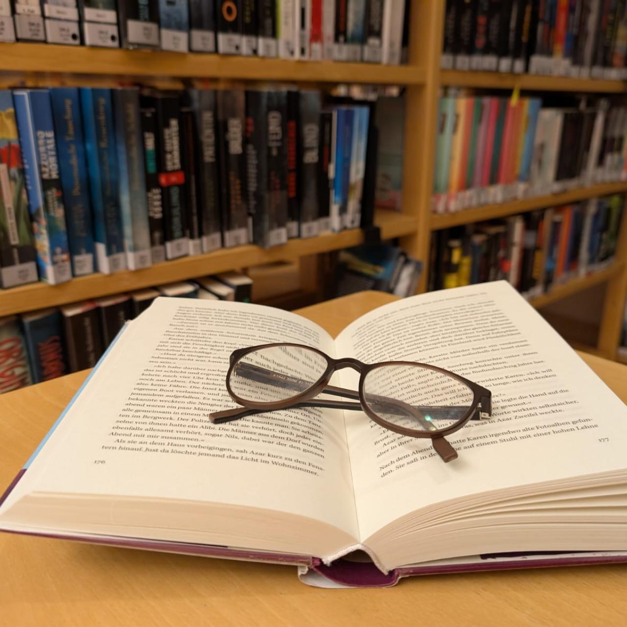 Ein Buch und eine Lesebrille auf einem Tisch, im Hintergrund ein Buchregal